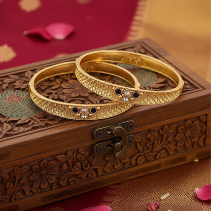 Two gold bangles with gemstones on a wooden box with floral patterns.