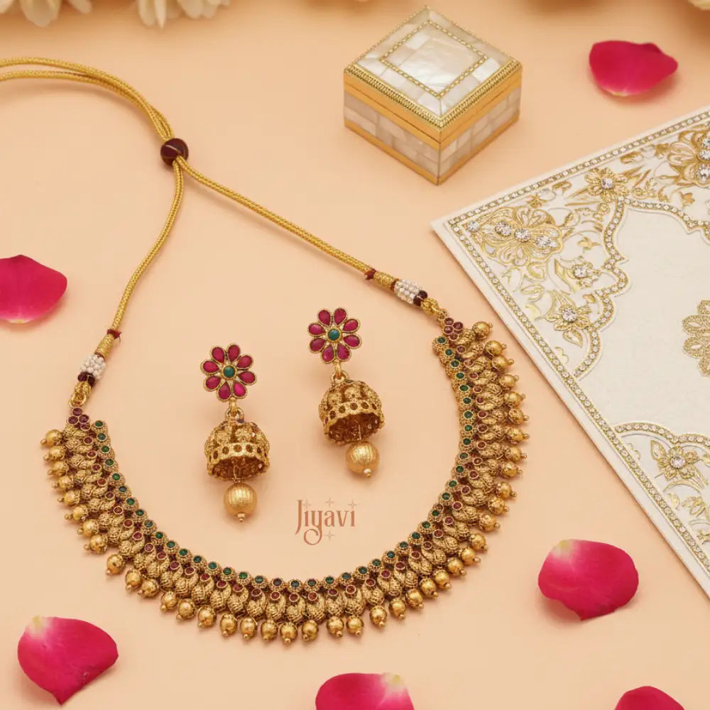 Gold necklace and earrings set with pink stones on a peach background, featuring the brand 'Jiyavi'.