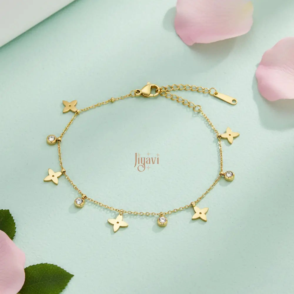 A delicate gold-plated chain bracelet adorned with several four-petal flower charms and small, sparkling clear stone on light green card paper, pink rose petals beside.