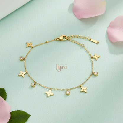 A delicate gold-plated chain bracelet adorned with several four-petal flower charms and small, sparkling clear stone on light green card paper, pink rose petals beside.