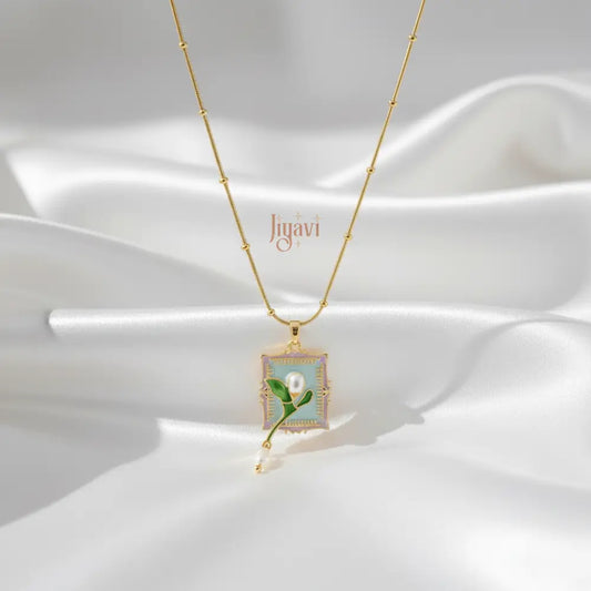 Gold necklace with a square pendant featuring a green leaf and pearl on a white fabric background.