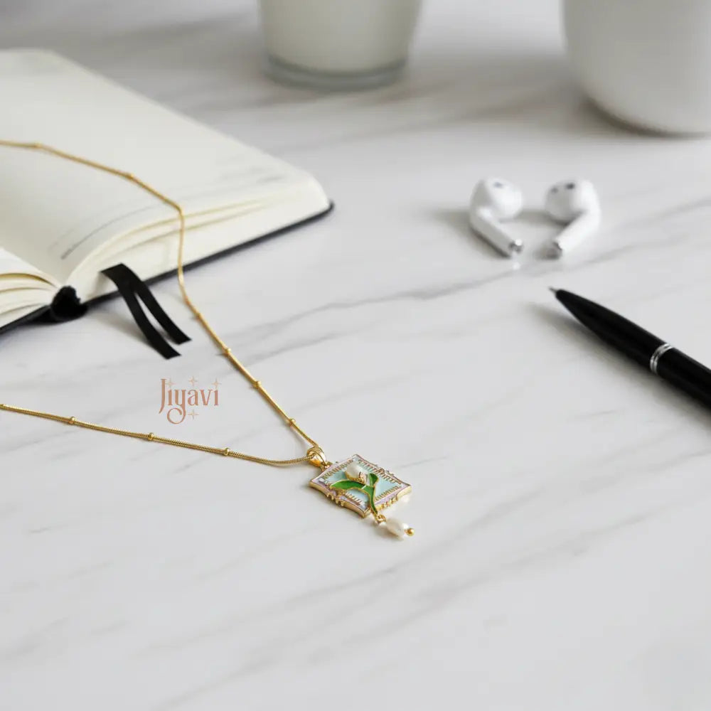 Gold necklace with a pendant on a white surface, next to an open book and a pen.