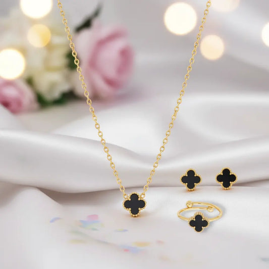Gold necklace and earrings with black clover pendants on a soft fabric background.