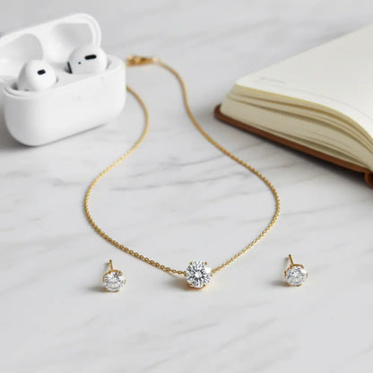 Gold necklace with diamond pendants and earrings on a light surface with an open book and white case.